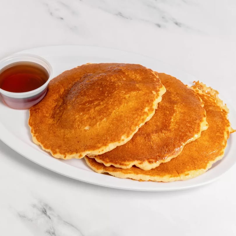 Three golden pancakes on a white plate with a side of maple syrup in a small plastic cup.