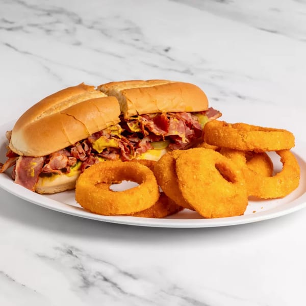 Two pastrami sandwiches with mustard and pickles, served with a side of crispy onion rings.